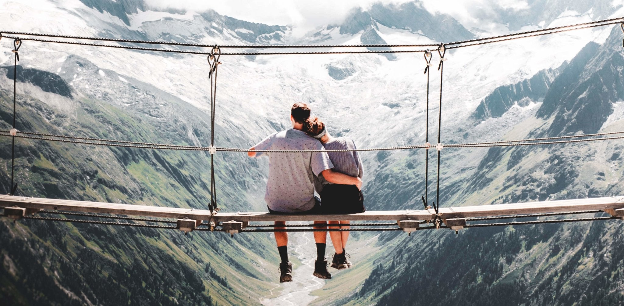 Two people sit on a suspended bridge overlooking snow-capped mountains and valley.