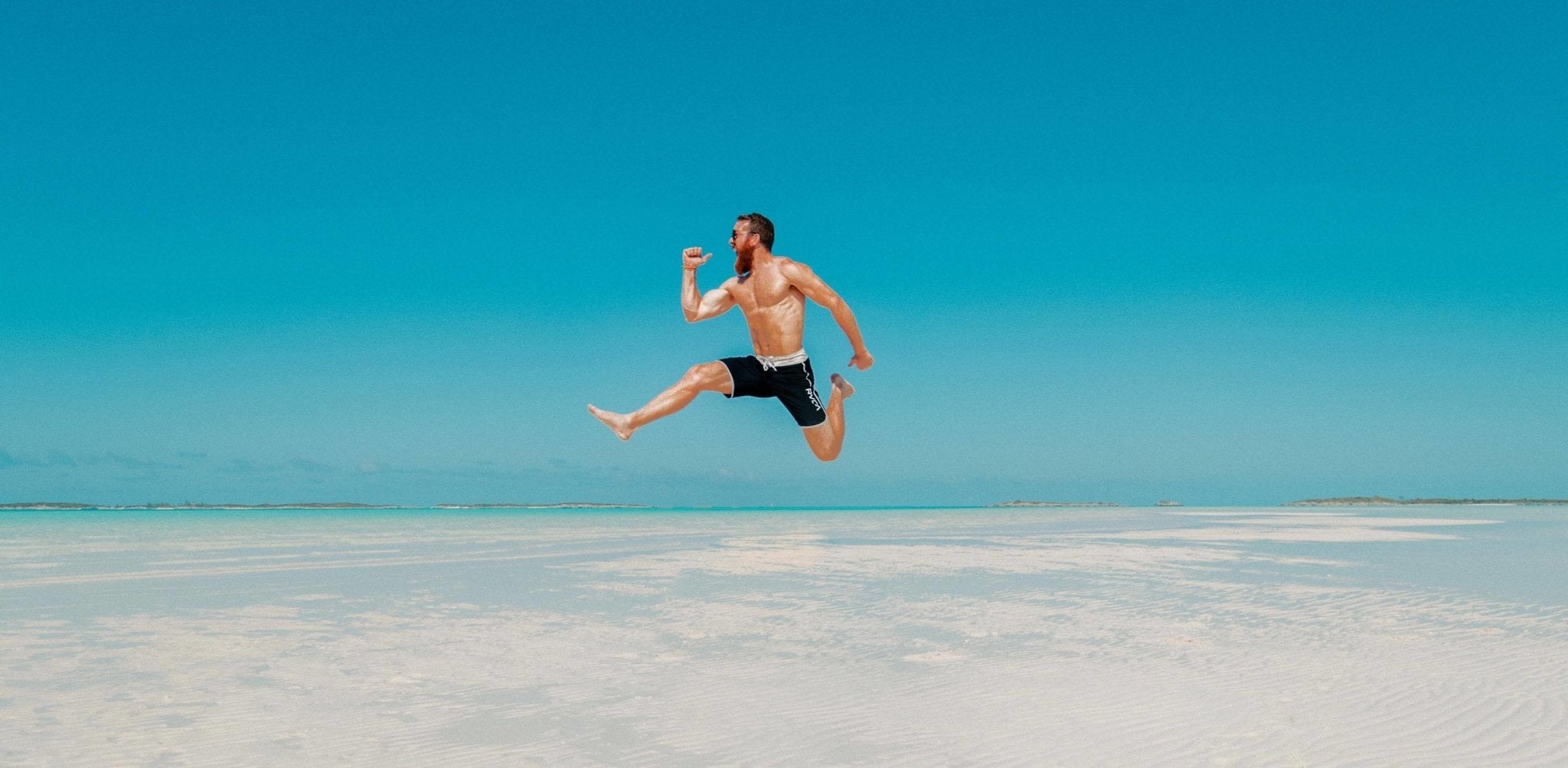 Shirtless man jumping on a beach with clear blue sky and shallow water.