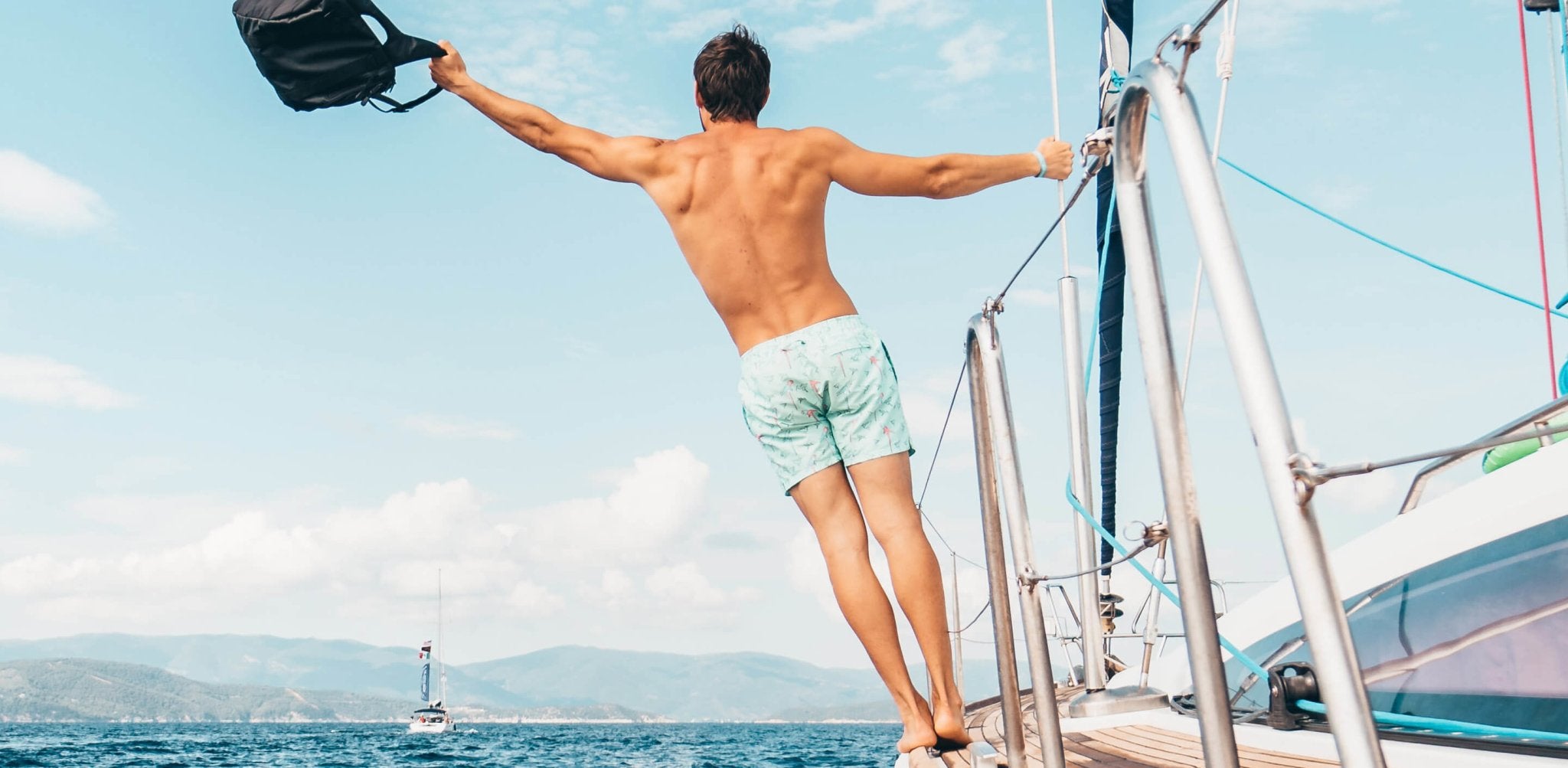 Shirtless man on sailboat with arms spread wide, holding hat, displaying bronzed back and shoulders.