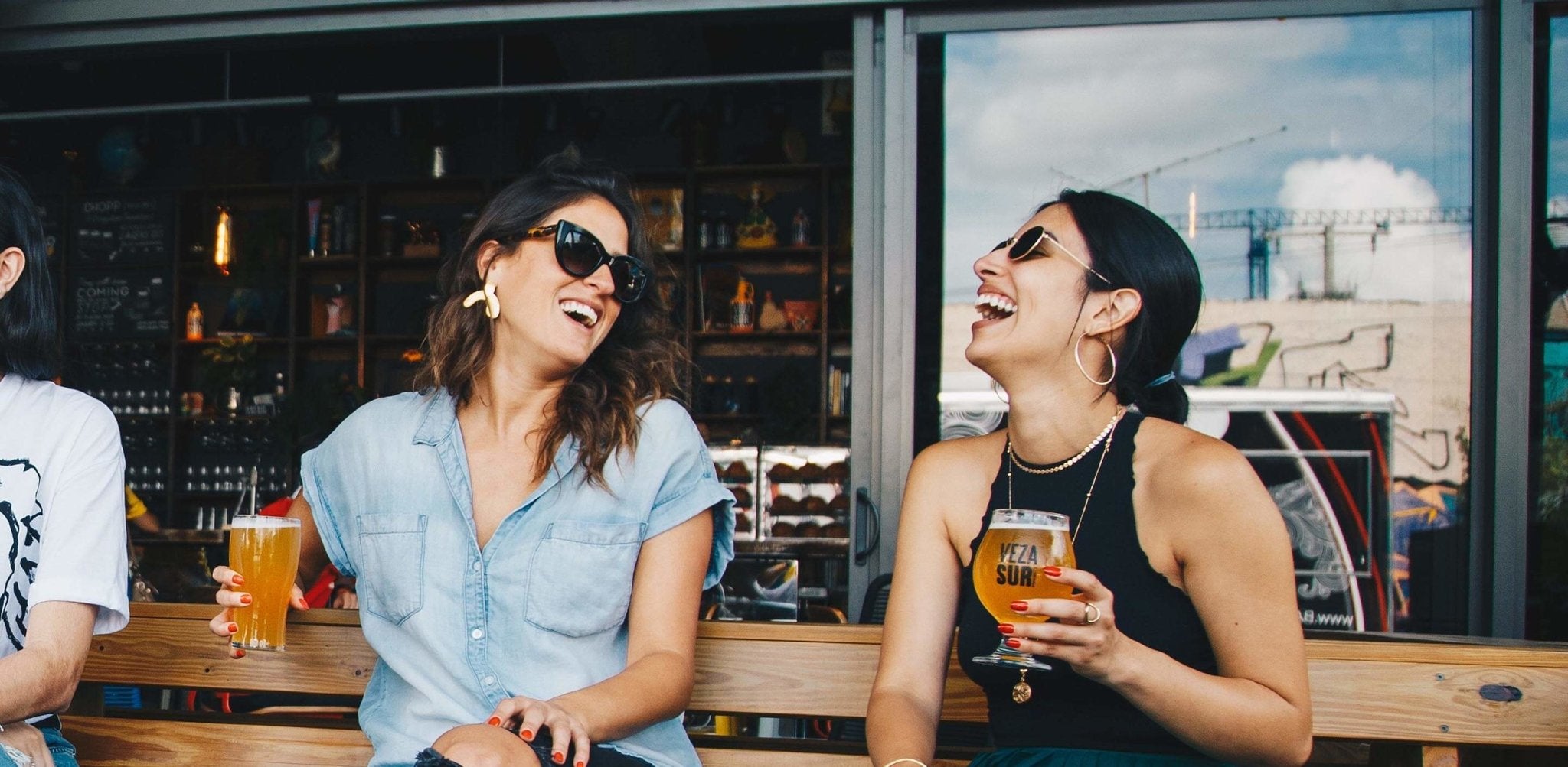 Two women laughing while holding drinks at an outdoor bar counter.
