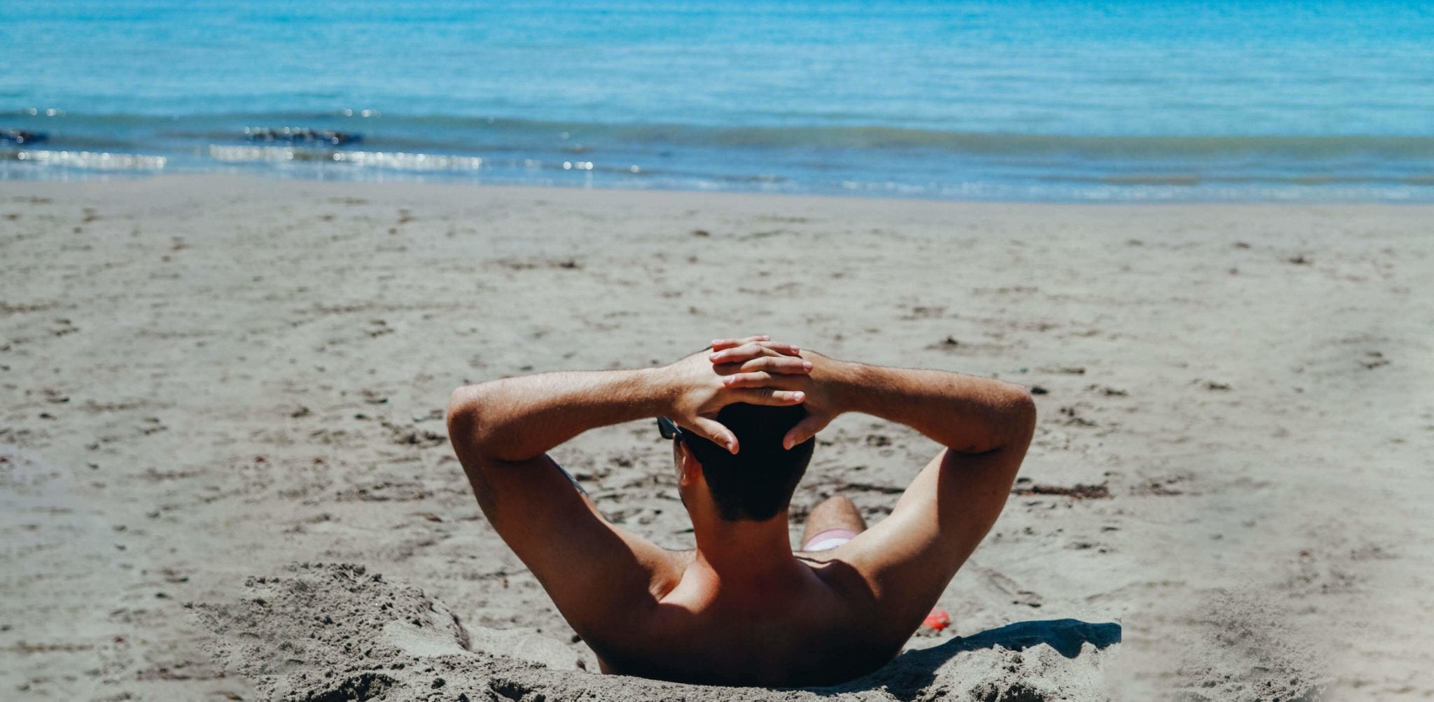Shirtless man relaxing on sandy beach with hands behind head, facing ocean.
