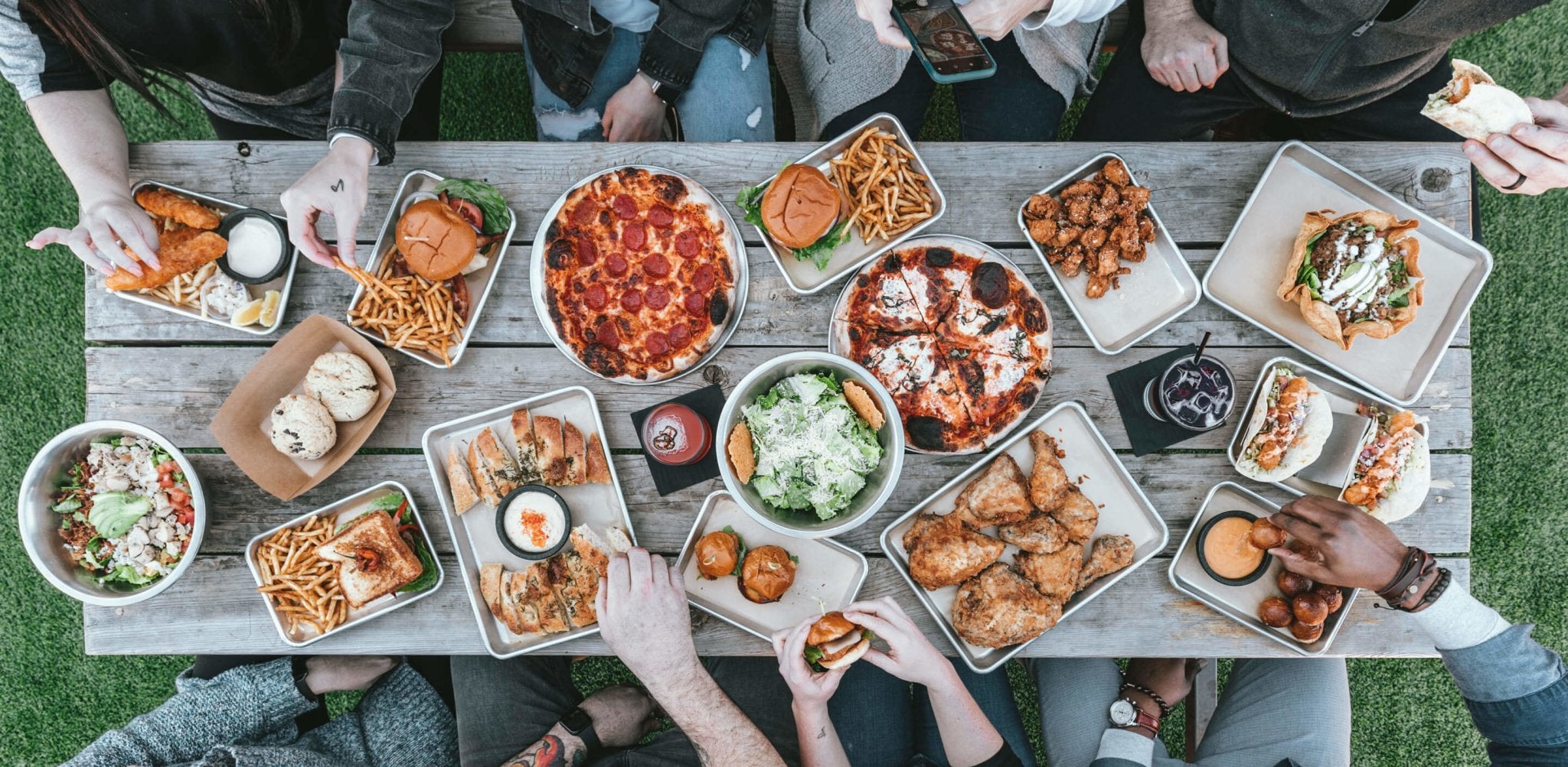 Overhead view of diverse foods including pizza, fried chicken, salads, and pastries shared among seated people.