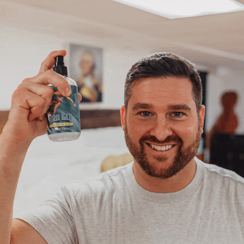 Man holding up a spray bottle with a dark label, smiling at the camera in a bright interior space.