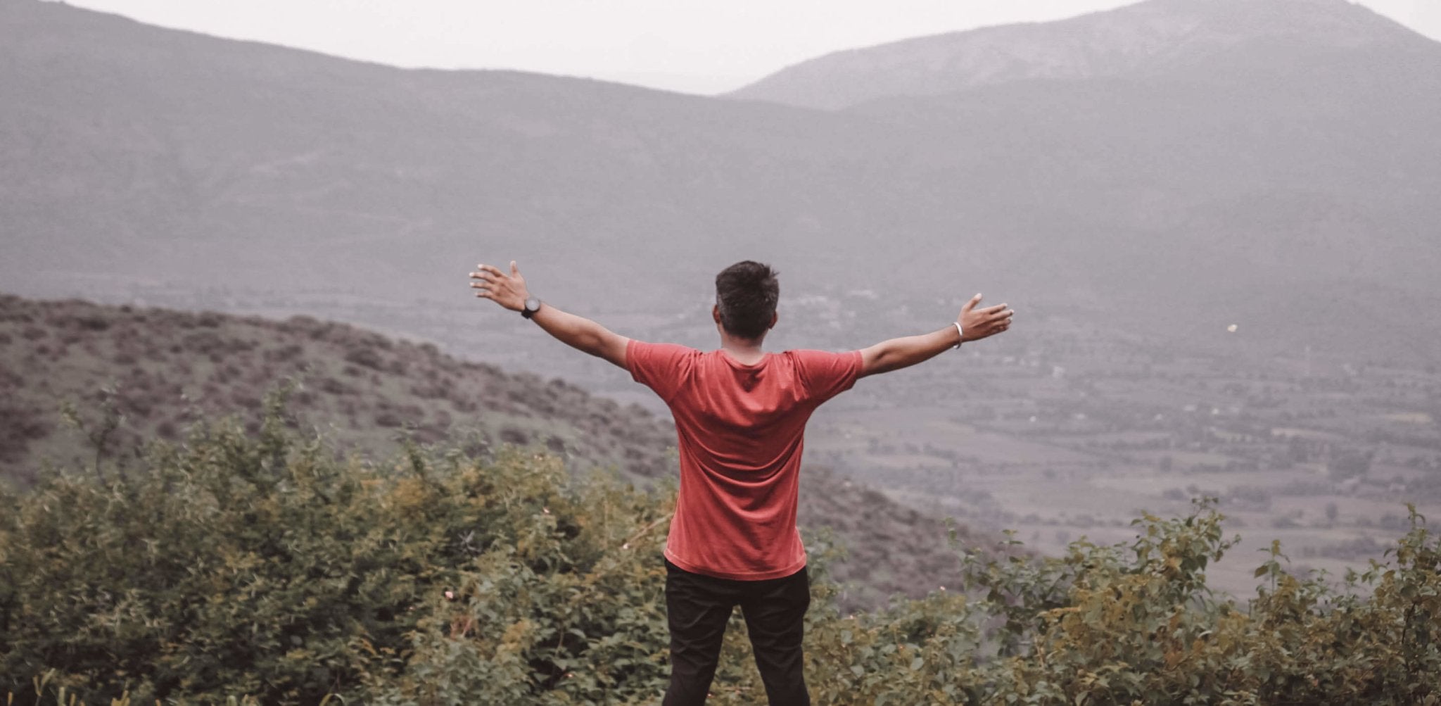 Man in red shirt standing with arms outstretched on mountainside, celebrating confidence and vitality.