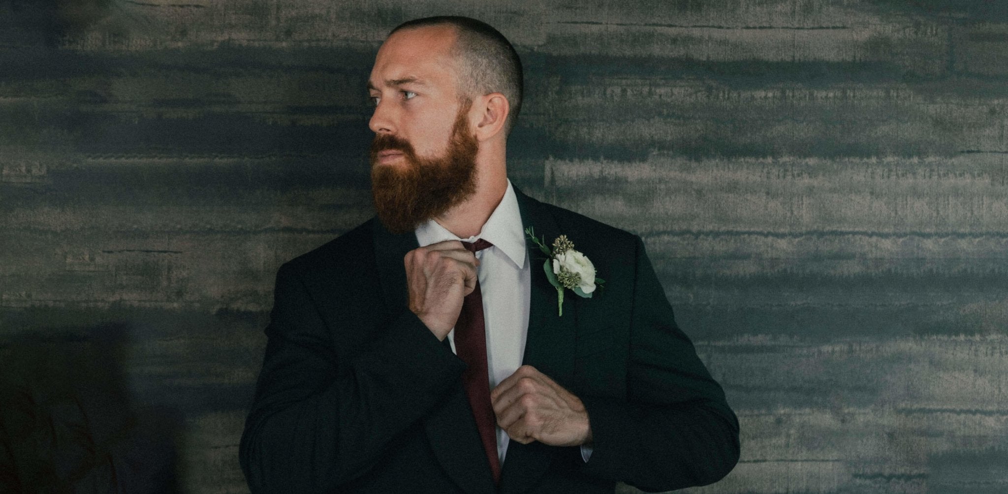 Bearded man in dark suit and tie adjusting his collar against textured backdrop.