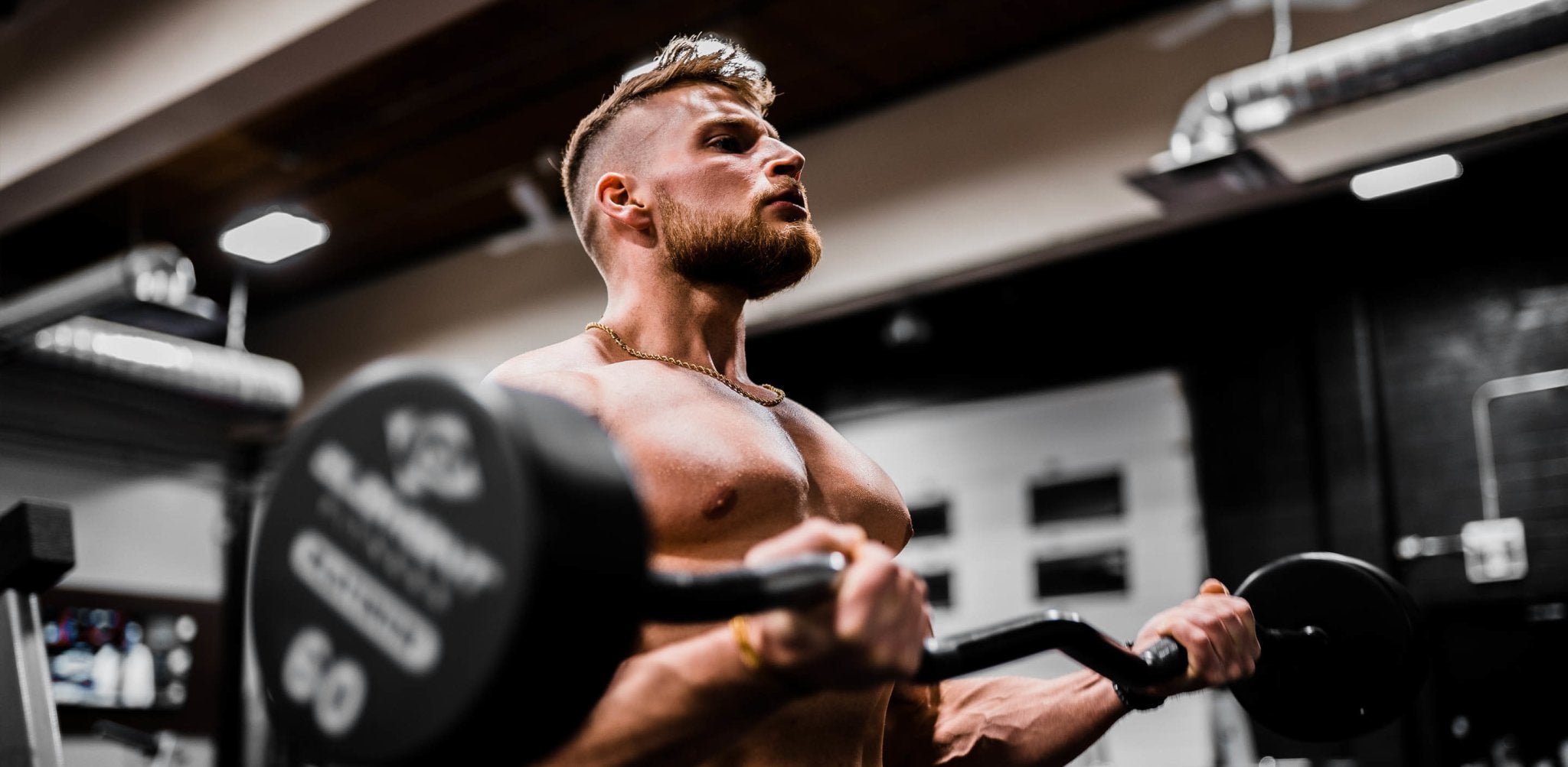 Muscular man in gym holding dumbbell, displaying tanned physique under studio lighting.