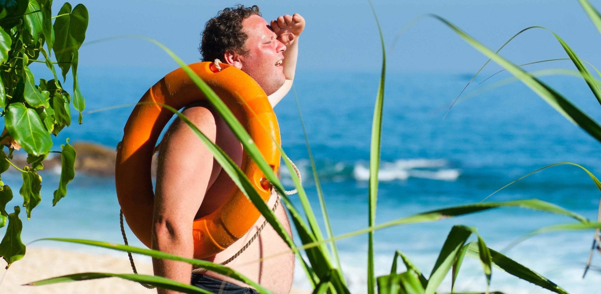 Man in orange life vest shielding eyes while looking at ocean under bright sunlight.
