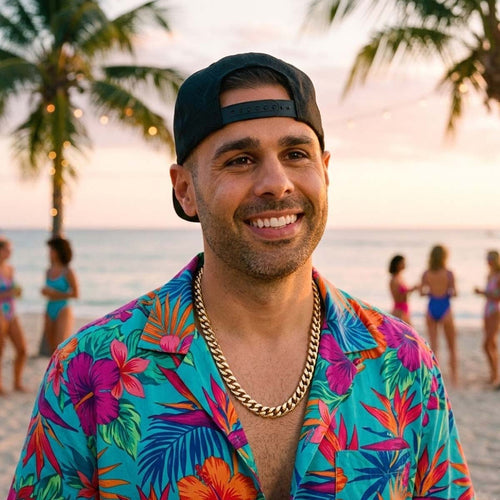 Smiling man in colorful tropical shirt and chain necklace at sunset beach with palm trees.