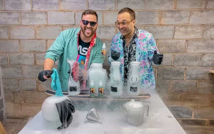 Two men spray-tanning each other against a brick wall, covered in white product and spray foam, laughing together.