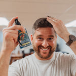 Man with beard spraying sea salt spray into his hair while smiling at the camera.
