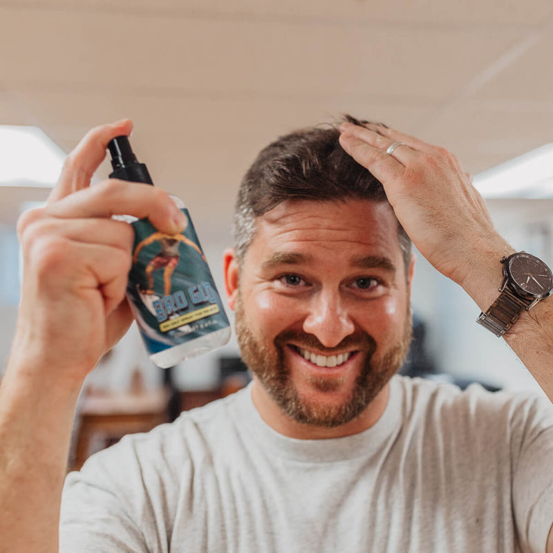 Man with beard spraying sea salt spray into his hair while smiling at the camera.