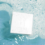 White exfoliating soap bar with embossed design soaking in turquoise soapy water with bubbles.