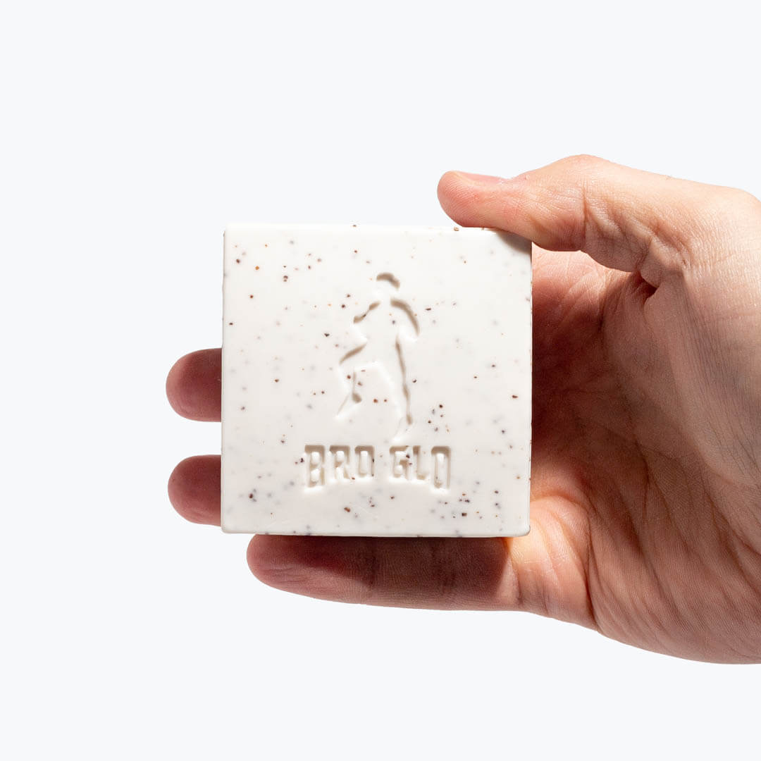 Hand holding a square exfoliating soap bar with embossed figure and speckled texture against white background.